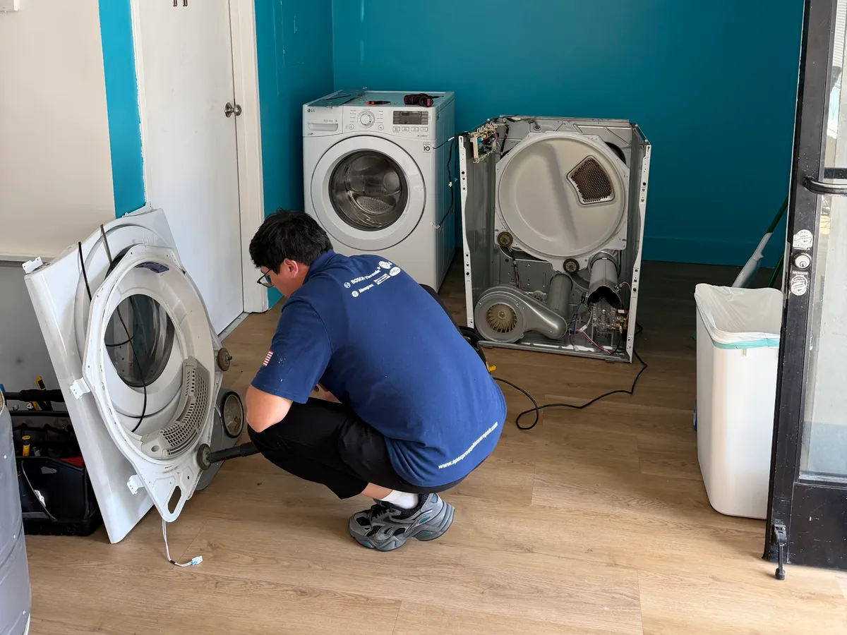 ApexPro technician crouched at the back of a dryer with the rear panel removed, drum and belt exposed, in the training bay