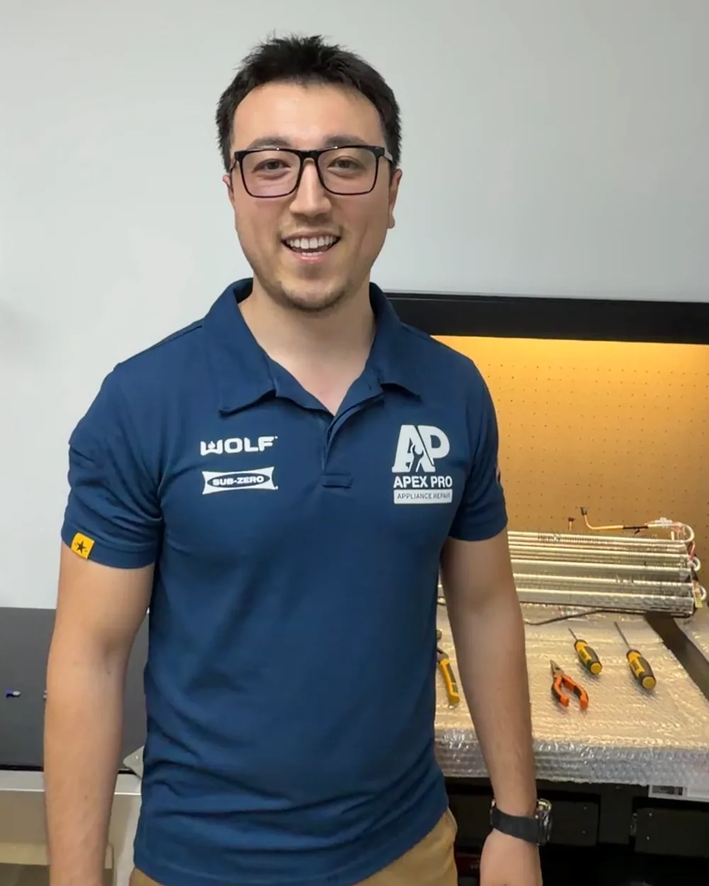 Joe, founder of ApexPro Appliance Repair, in a navy branded polo standing beside a refrigerator evaporator coil on a workshop bench